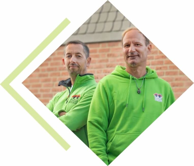 Porträtfoto von den Geschäftsführern. Beide tragen einen grünen Firmen-Pullover und stehen leicht angelehnt zu einander.